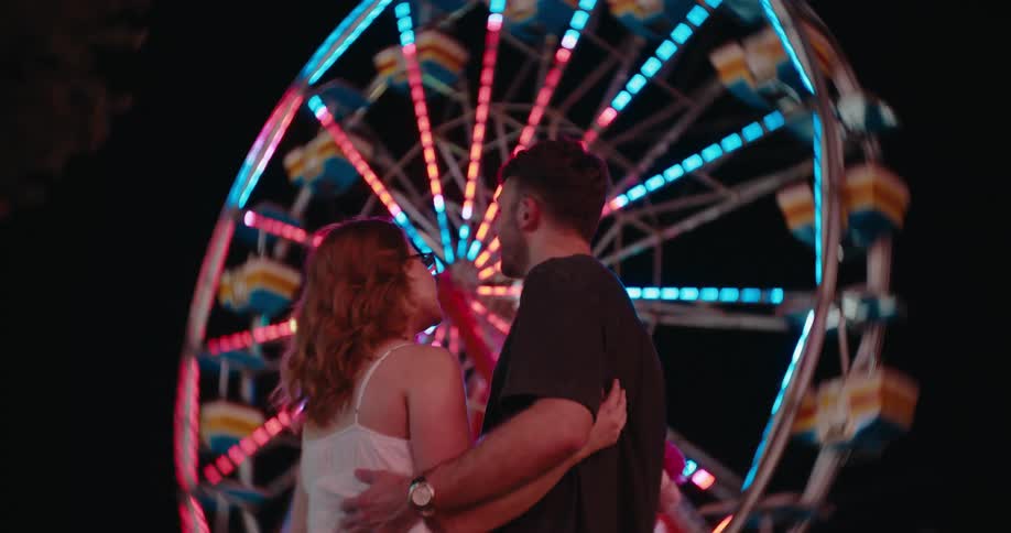 A man and a woman stand facing each other and embracing in the foreground with the background dominated by a large rotating wheel ride illuminated by multiple rows of changing colored lights against a dark night sky