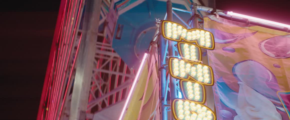 A bright yellow carnival sign at night with the illuminated multicolor structure of a ferris wheel behind it