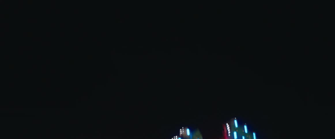 An amusement park ride covered in rows of brightly colored lights rotates and swings its arms in complex patterns against a dark night sky with the light trails creating visual streaks of color