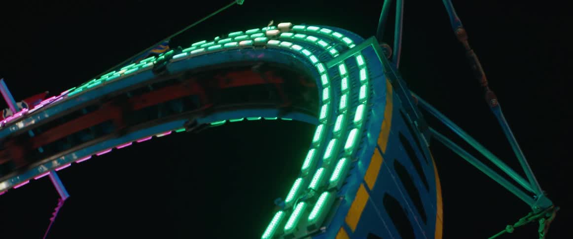 A moving amusement park ride shows a blue and yellow structure rotating rapidly against a dark background with rows of colorchanging lights illuminating the curved track and riders visible as the vehicle passes