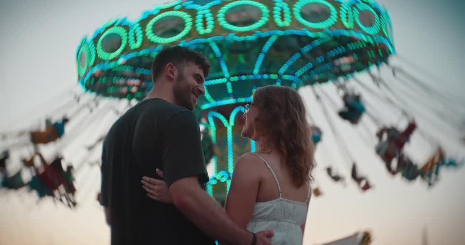 A man and a woman stand close to each other with the mans arm around the womans shoulder as they face toward a large brightly lit swing ride that rotates rapidly in the immediate background against an overcast sky