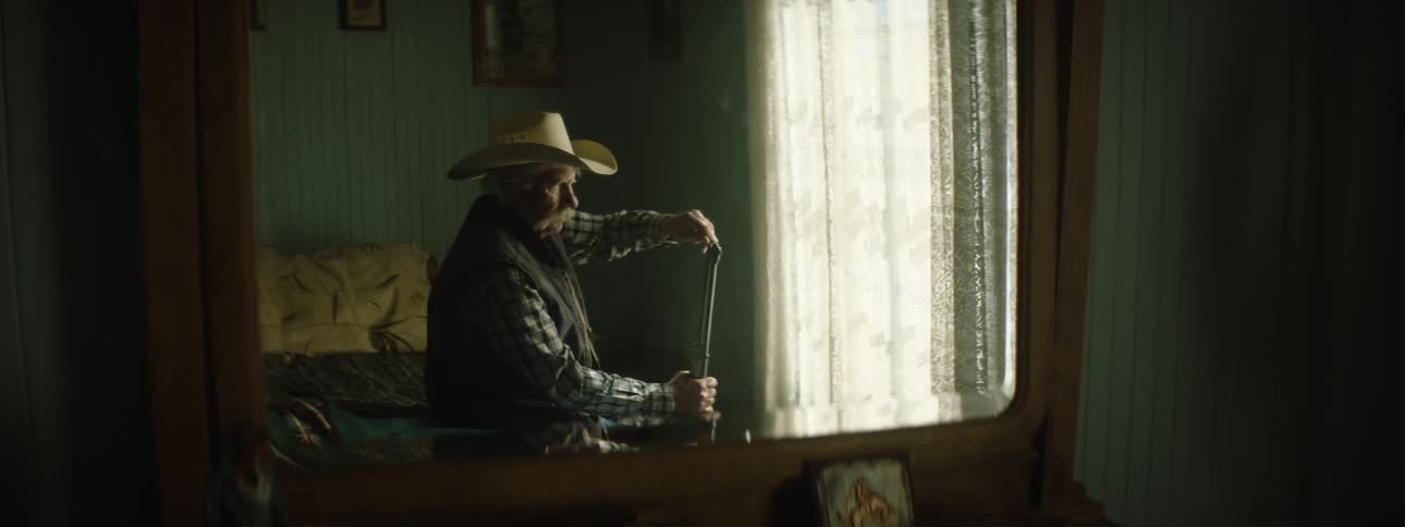 An elderly cowboy holds a gun on a bed