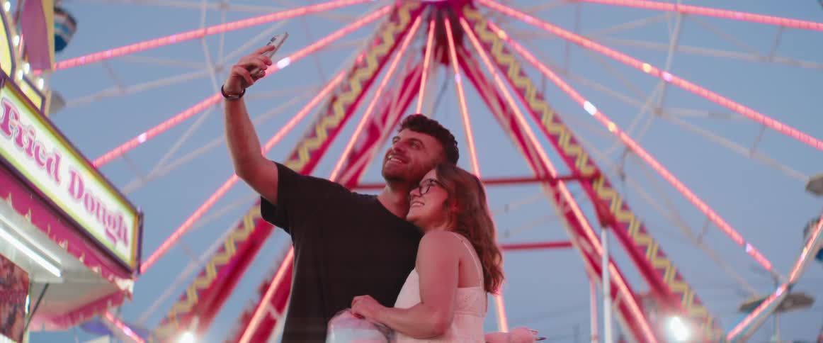 Couple taking a selfie on their smartphone and smiling at a fair outdoors during night time