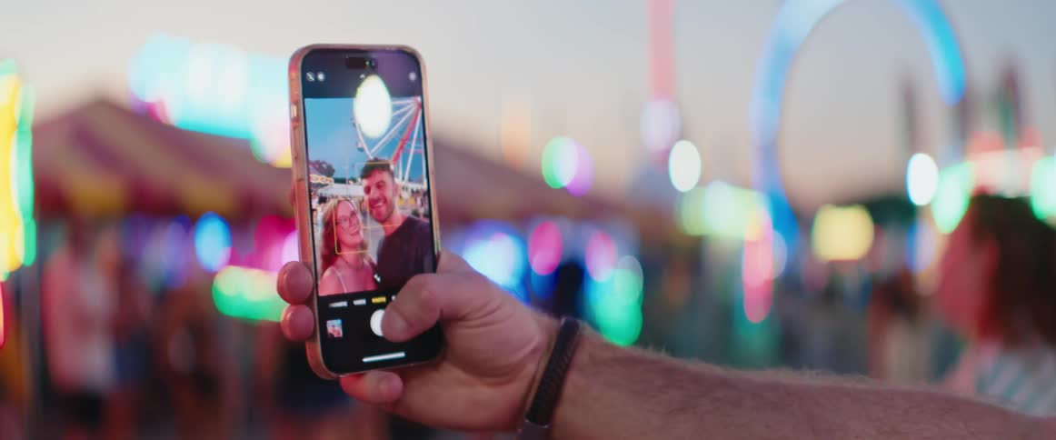 A persons hand holds a mobile phone vertically in a closeup with the phone screen displaying the live camera view of a man and a woman posing together in an amusement park blurried