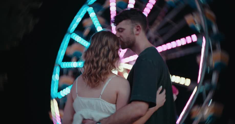 Young male and female hugging and kissing in front of a ferris wheel outdoors during night time