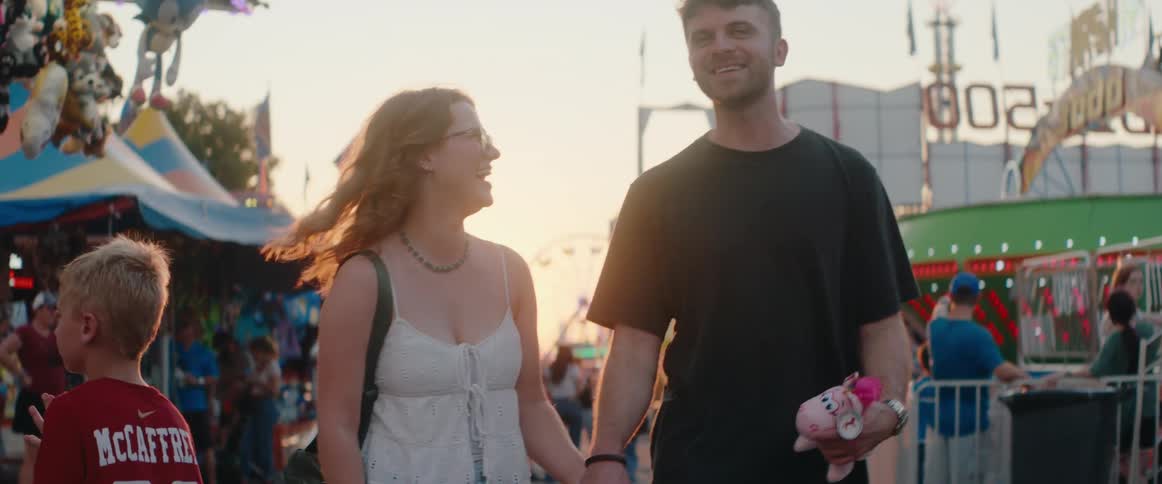 Couple walks hand in hand smiling at a fair during sunset