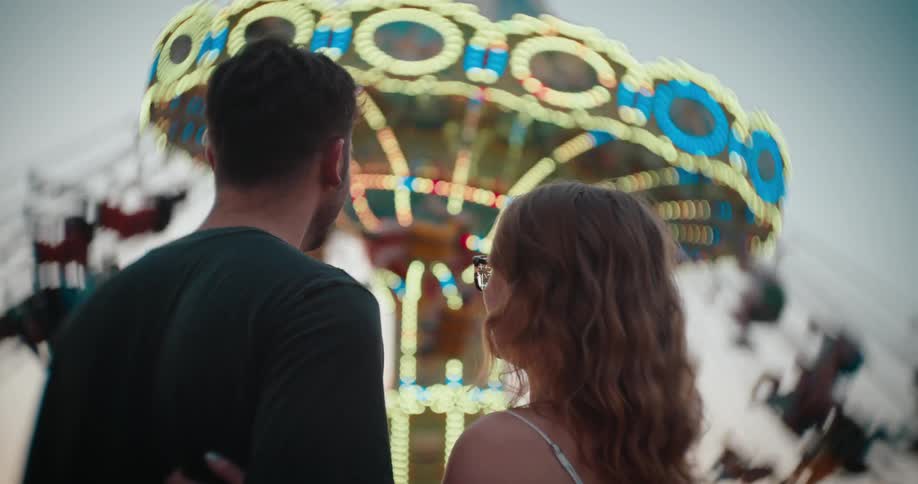 A man and a woman standing face to face and smiling at each other with a brightly lit rotating amusement park ride structure out of focus in the background