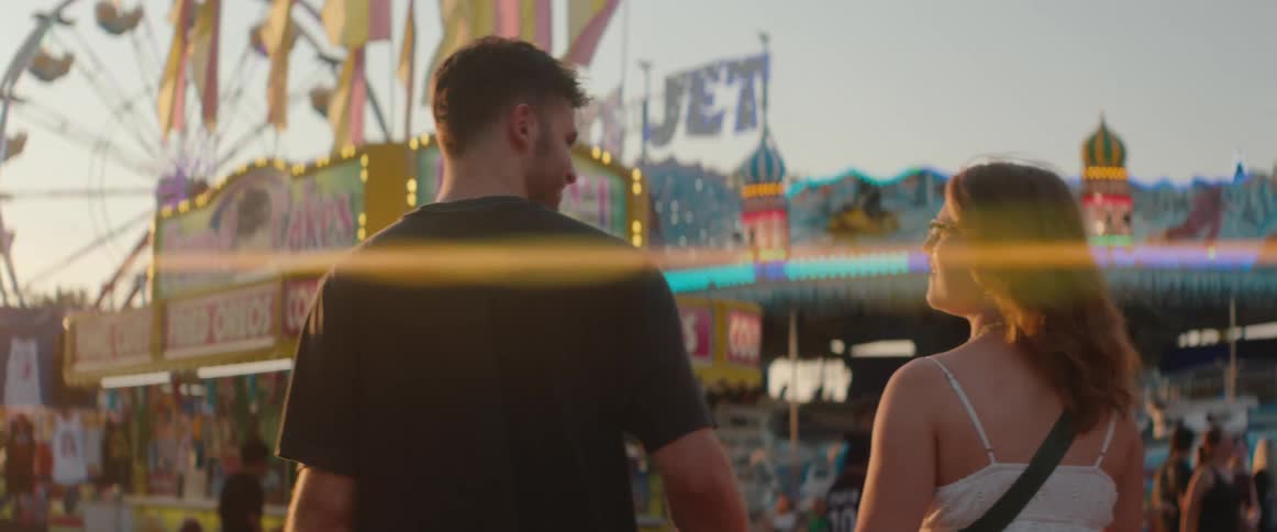 A man and a woman walk through a sunny carnival as a lens flare streaks across a ferris wheel and food stands behind them