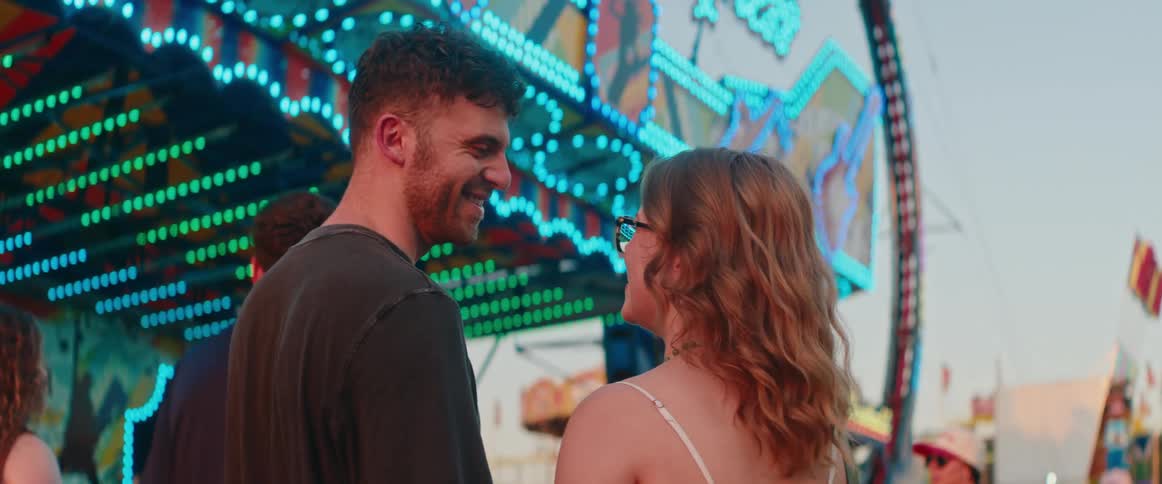 A smiling man and woman talk at a brightly lit carnival ride during a bright day