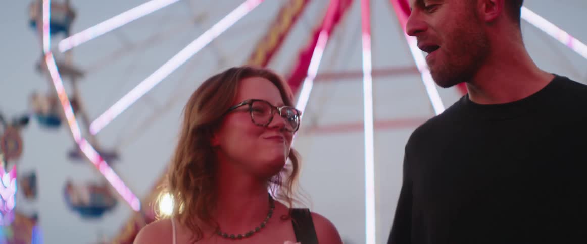 Couple enjoys eating cotton candy in front of a brightly lit ferris wheel