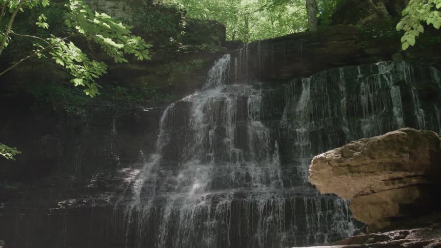 Water flows in multiple vertical streams down a wide dark tiered rock face surrounded by dense green vegetation with a large lighter rock formation resting in the right foreground under diffused sunlight