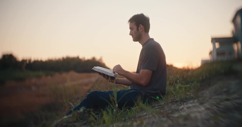 A man sits on a grassy mound facing toward the left holding an open white book in his lap and looking down at it while a blurred treeline and distant residential structures sit beneath an orange and yellow sky