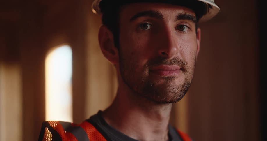 A medium eye level view of a bearded man wearing a white hardhat and an orange safety vest looks directly at the camera inside the wooden frame of a construction site