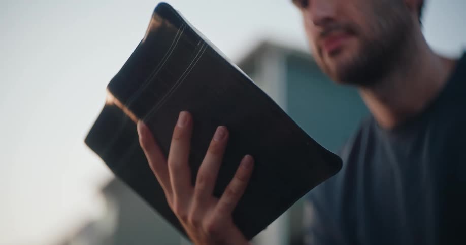 A man holds a leather bound book possibly a bible and reads intently outdoors as the sun sets