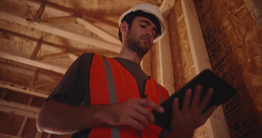 A low angle shot shows a construction worker wearing a white hardhat and an orange safety vest inspecting a wooden ceiling structure while reviewing plans on a tablet computer