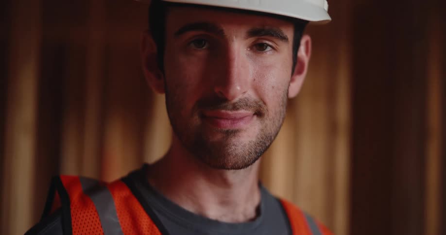 A close head and shoulders view shows a smiling bearded man wearing a white hardhat and an orange safety vest looking up to smile directly at the camera inside a wooden structure