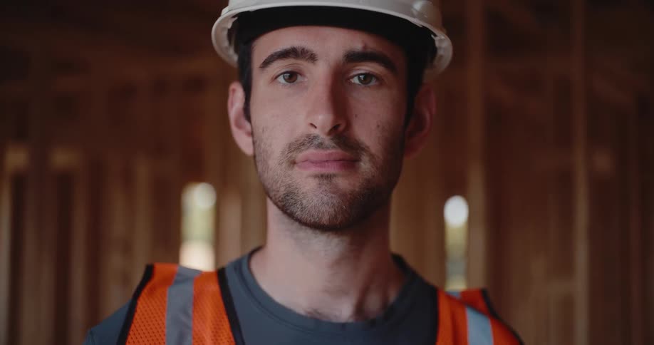 A closeup shot of a man wearing a white helmet and an orange reflective vest looks down then slowly raises his eyes to look directly at the camera while standing inside the wooden framework of a structure
