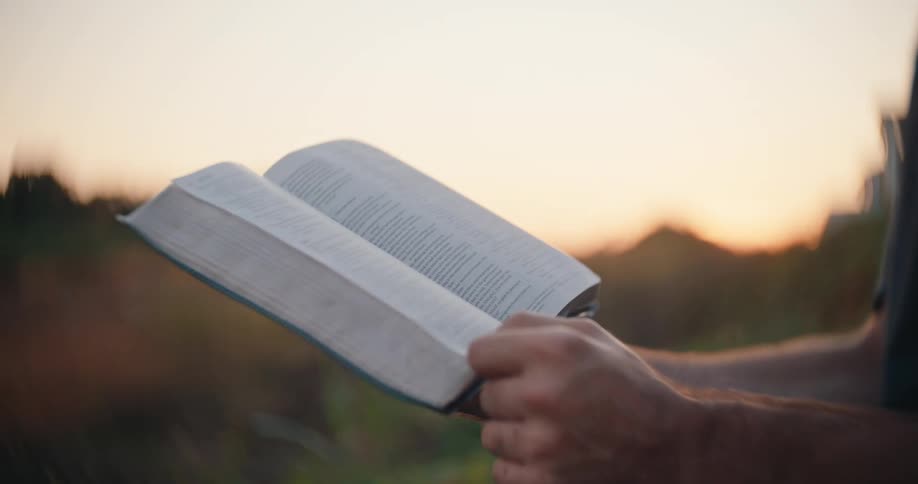 A persons hands holds an open bible at chest level and turn a page against a blurred background and a sky showing the soft orange and yellow light of a setting sun