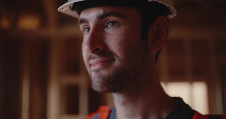 A construction worker in a hard hat and safety vest looks upward inside a woodenframed building with a focused thoughtful expression