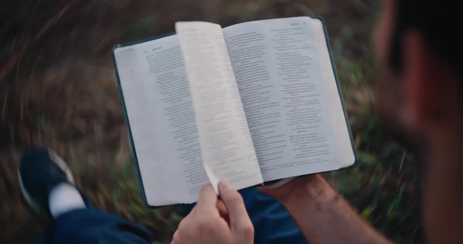 A man sits outdoors in the grass holding an open bible and gently lays his hand flat on the pages before turning one in an overhead close view