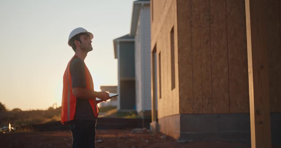 A man wearing a white helmet and an orange reflective vest stands outside on a dirt surface between a partially constructed building and another house looking up at the structure while holding a tablet in his hands