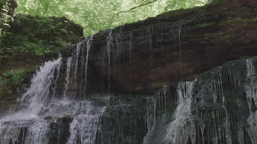 A wide sheet of water flows over the edge of a high multitiered dark rock cliff covered with moss then breaks into many individual vertical streams as it descends with dense green tree cover visible above the rock face