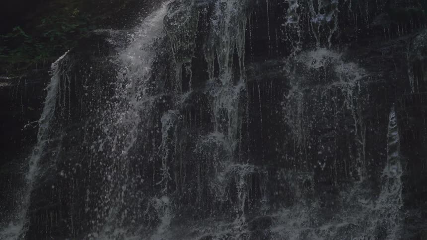 Water flows in many thin and thick streams down a wide steep dark and irregularly stepped rock face creating a shimmering curtain effect against a shadowed natural background