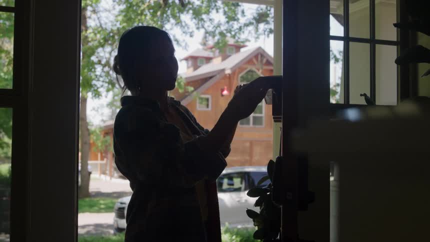 A woman stands framed in a doorway silhouetted against a bright outdoor scene showing a wooden building and uses her hands to operate a mechanism on the side of the doorframe