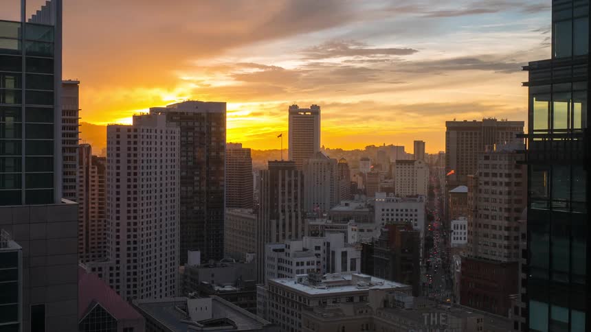A dramatic aerial time lapse view shows a cityscape of tall buildings darkening as the sun sets casting an intense yellow and orange glow across the sky and clouds