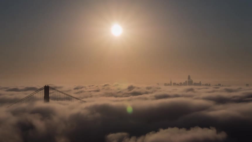 The bright sun hangs in a hazy sky above a sea of clouds with the top of the golden gate bridge and the san francisco skyline visible in the distance