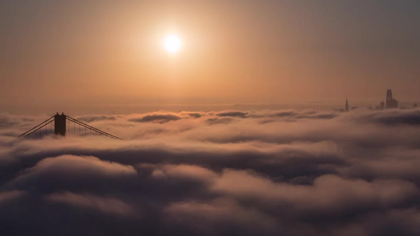 Sun rises over a city partially covered by clouds revealing a bridge tower