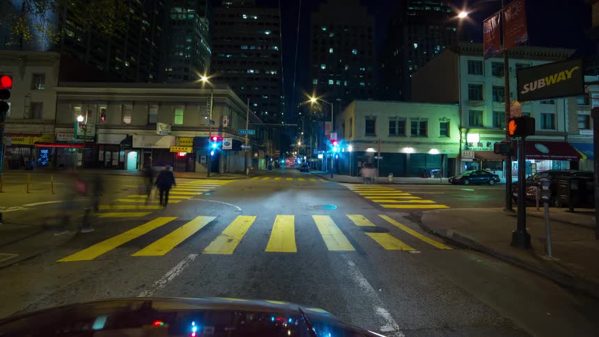 A dramatic time lapse and motion blur shot from a moving car captures the bright streaks of headlights and city lights at night as the vehicle drives quickly through a busy skyscraper lined street