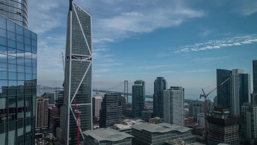 A dense urban skyline surrounded by numerous other skyscrapers with a bridge visible in the distance over a large body of water under a partly cloudy blue sky