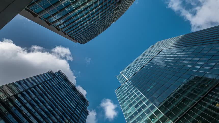 A view looking up at modern glass skyscrapers reflecting clouds against a bright blue sky