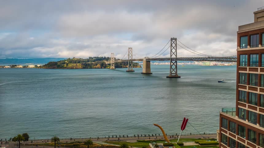 A wide view of a large suspension bridge spanning a body of water is framed by a modern brown multistory building on the right and a walkway with landscaping in the foreground as a small vessel moves across the water under a sky with scattered clouds