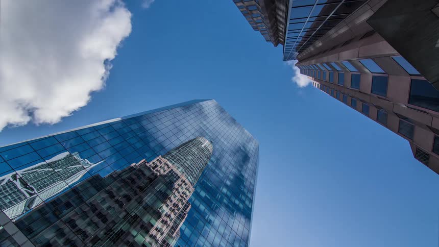 A lowangle timelapse shows clouds speeding across the blue sky their movement mirrored in the reflective glass of modern skyscrapers