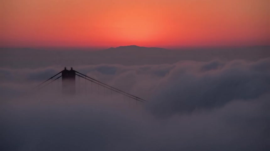 A suspension bridges towers rise through thick coastal fog at sunrise as distant hills silhouette against an orange sky