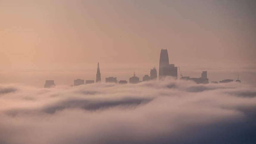 The dark silhouette of a group of tall urban buildings including a prominent tapered tower and a flattopped skyscraper rises out of a dense layer of moving white fog or low cloud that extends across the entire foreground under a sky colored pale orange