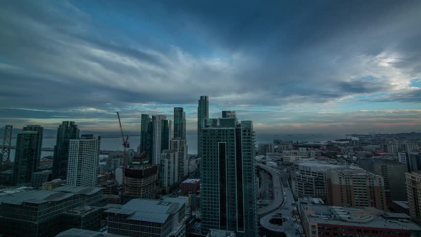 A high angle urban time lapse shows dark clouds quickly crossing the sky above a dense cityscape of tall modern skyscrapers and highways leading to a distant bay bridge