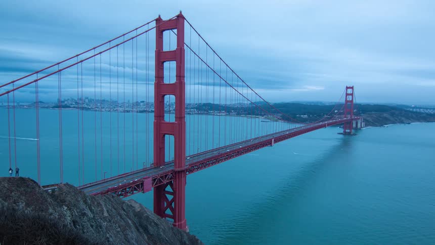 Traffic crosses a suspension bridge while watercraft pass under it as night becomes day