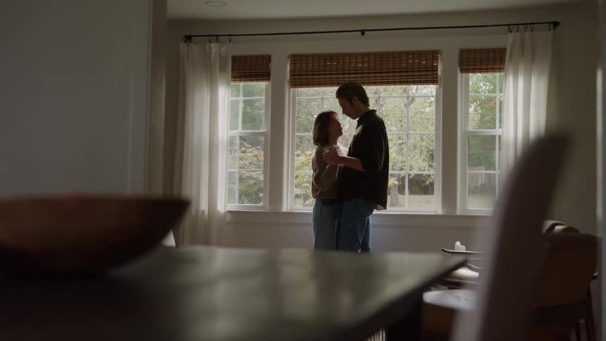 A man and a woman stand close together facing one another in front of a threepane window with white curtains and woven blinds silhouetted against the bright daylight from outside while blurred furniture occupies the foreground