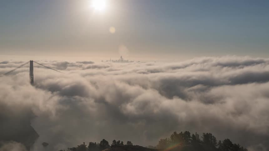 Thick fog rolls over the golden gate bridge with the san francisco skyline faintly visible in the distance under a bright sun