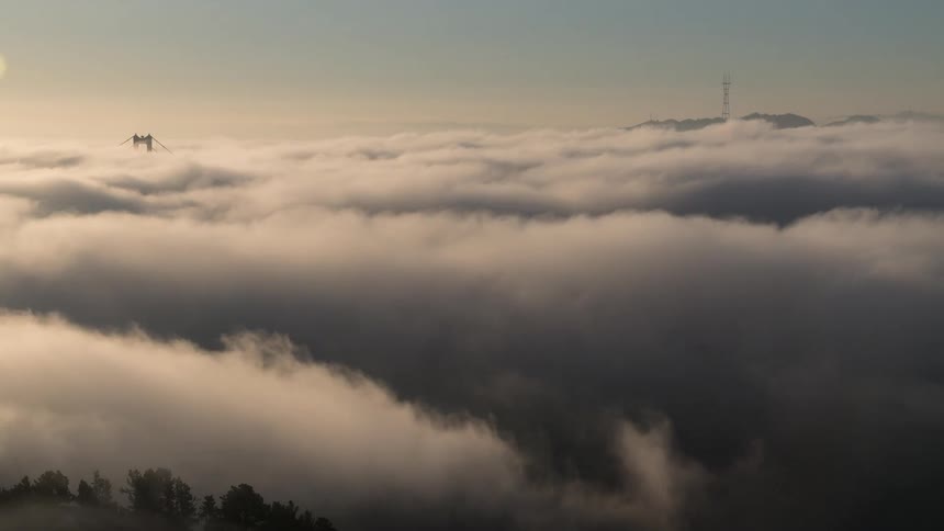 A sea of morning fog blankets hills with only distant towers and bridge peaks poking through under a pale sky