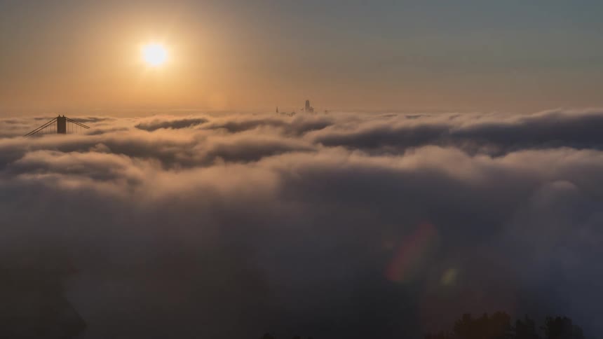 A dramatic aerial time lapse shows the golden gate bridge and a city skyline peaking out from a massive expanse of rolling low fog as the bright morning sun shines above