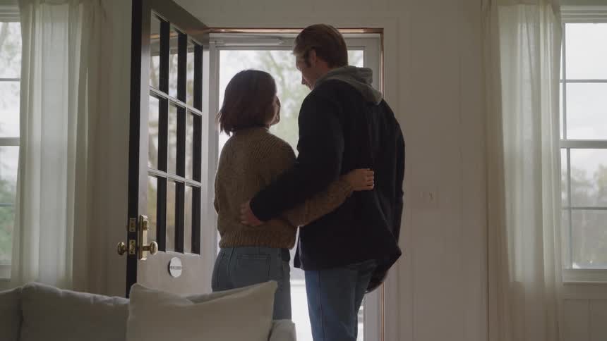 A medium shot of a man and a woman embracing and kissing in a doorway before the man puts on a gray hoodie and a dark jacket grabs a white hardhat and walks out the door