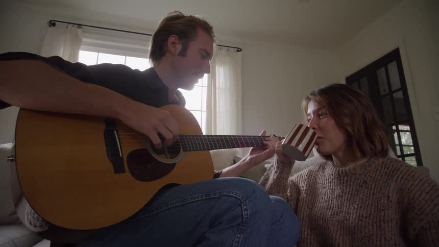 A man plays an acoustic guitar on a couch while a woman in a chunky sweater sits on the floor beside him smiling as she sips from a striped mug in a cozy living room