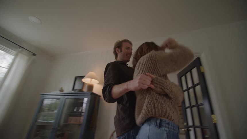 A smiling man in a dark shirt and a woman in a brown sweater and jeans playfully twirl and dance embracing each other in the brightly lit living room of a cozy home