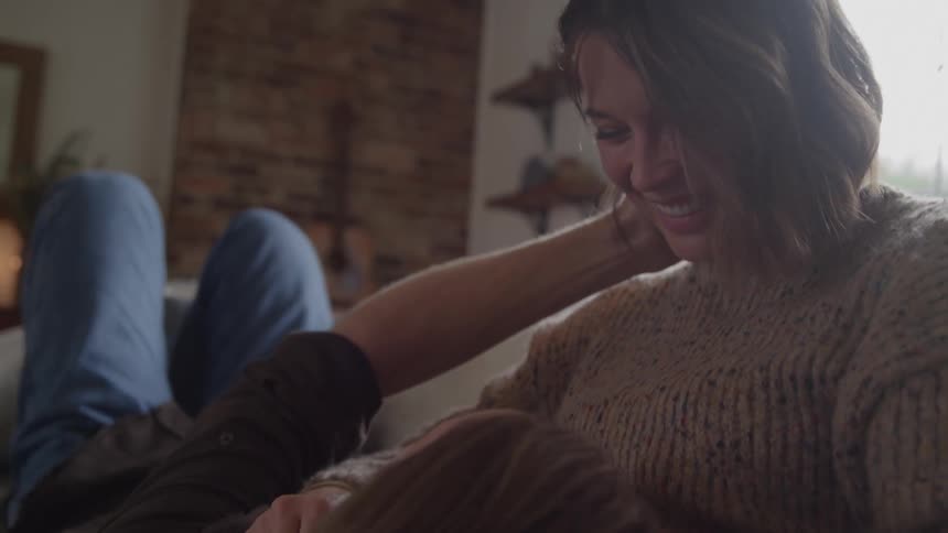 A woman in a chunky knit sweater cradles a mans head in her lap on a couch gently stroking his hair in a softly lit living room