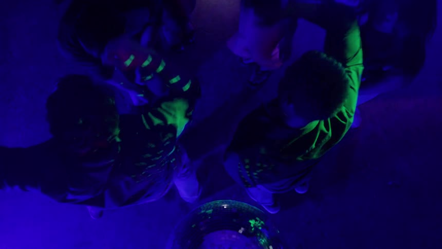 An abstract high angle shot of five young people dancing around a green disco ball on the floor of a dark club as flashing red and blue lights illuminate the scene