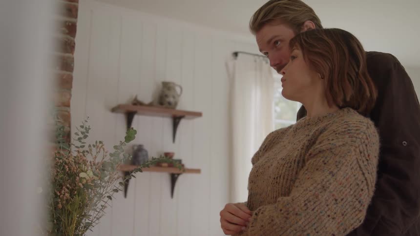 A woman wearing a knit sweater stands facing a wall then turns her head slightly as a man in a dark shirt moves close to her from behind and embraces her in a domestic setting